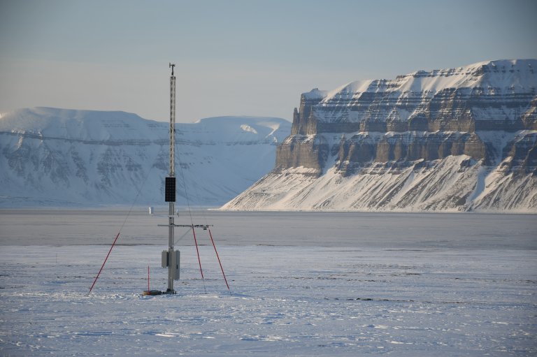 En høy måler står plassert i et hvitt landskap av snø og is, fjell i bakgrunnen. 