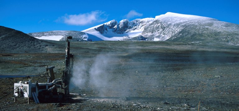 En boremaskin står plantet midt i fjell-landskapet på Dovre. Blå himmel og snødekket fjell i horisonten.